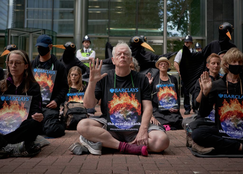 People dressed in black, sit meditating in front of a bank. They are wearing placards which say 'Barclays: the ecocide bank' and show a world on fire.  The meditators sit with their eyes closed but with one hand raised in a mudra. 