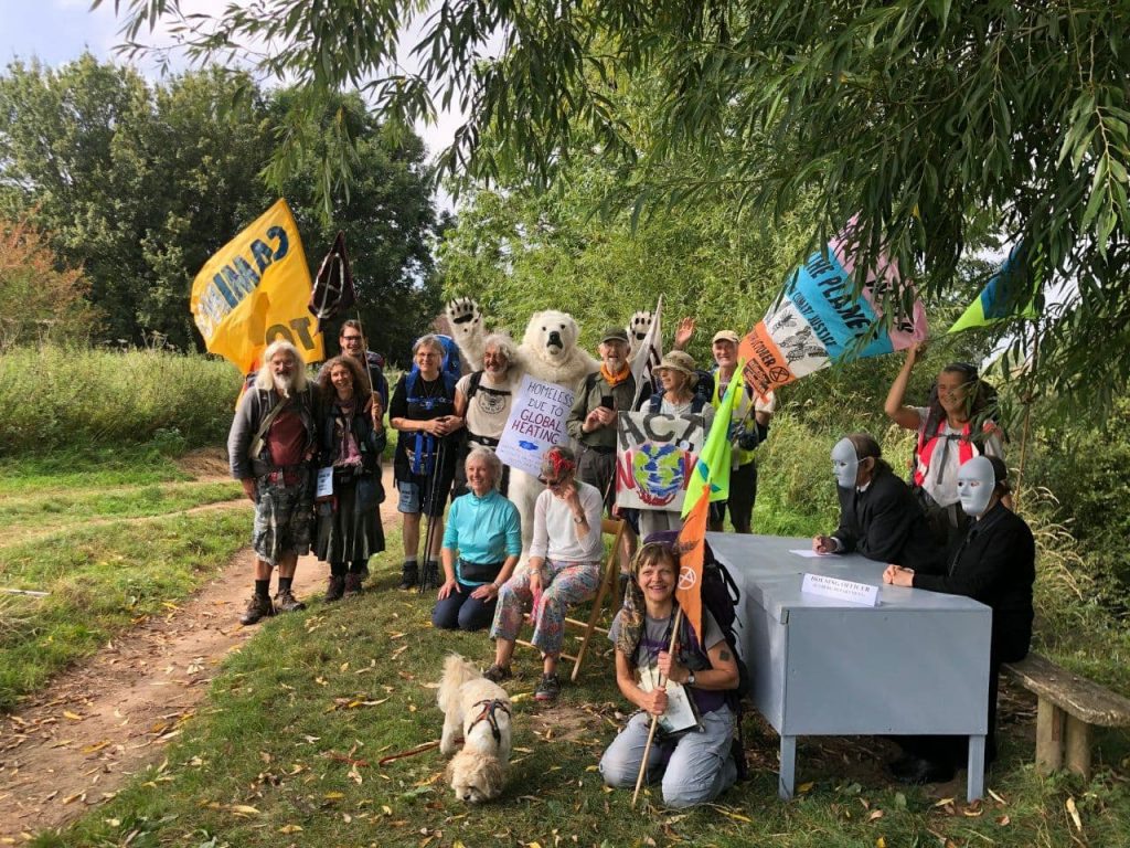 a group of people stand together smiling and waving . They are in a field, underneath a willow tree. They are carrying flags and one of them is dressed as a polar bea.