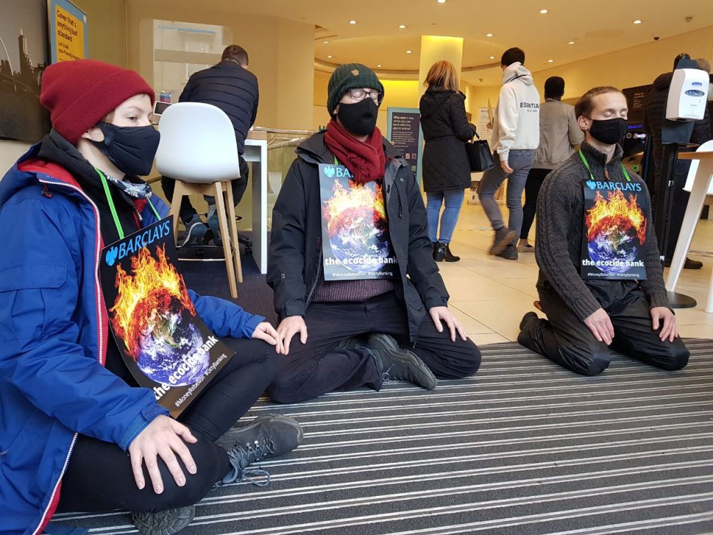 Three people sit in meditation wearing placards  that read: Barclays the Ecocide Bank. 
