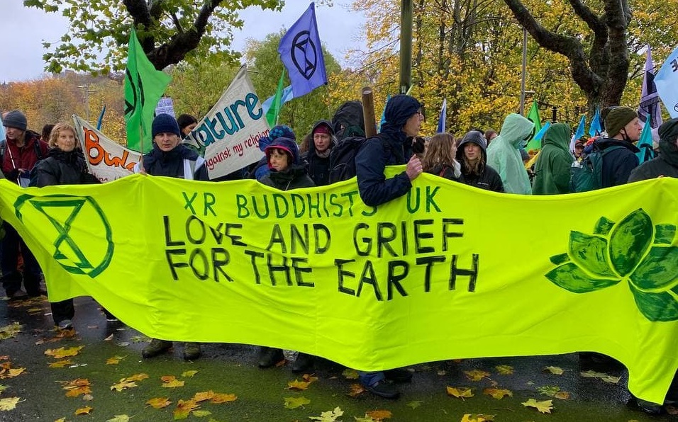 Protesters standing in the rain, carrying a bright yellow/green banner which reads 'XR Buddhists UK Love and Grief for the Earth'