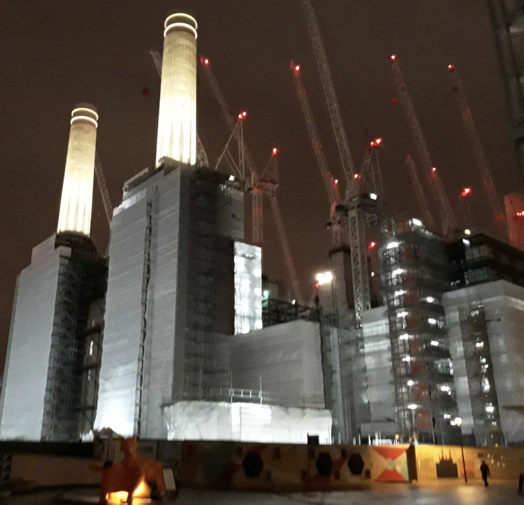 Battersea PowerStation at night, high grey buildings in the foreground topped by brightly illuminated tall chimneys. In the background, a collection of tall cranes.